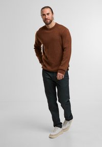 Pull en tricot marron avec col rond, pantalon noir et baskets blanches. Texture lisse, coupe décontractée et détails subtils sur les manches.