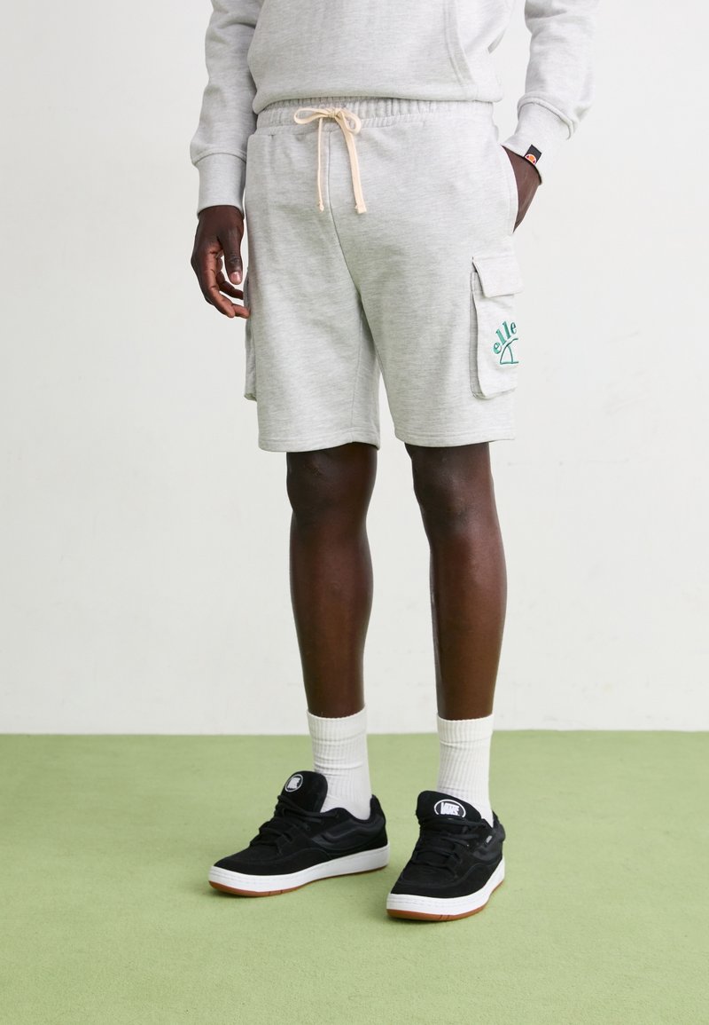 Harmaat collegehousut, joissa on naruvyötärö, sivu cargo-taskut ja vihreä logo. Yhdistetty mustiin lenkkareihin ja valkoisiin nilkkasukkad.