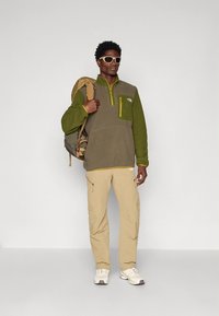 Flis pulover v olive zeleni barvi z rumenim zadrgo in žepom, kombiniran z bež hlačami in belimi supergami, spremlja ga sivi nahrbtnik.