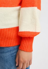 Pleteni pulover z oranžnimi in kremnimi vodoravnimi črtami. Riblji manšeti in rob. Mehak material, sproščen kroj. Roka resting na modrem džinsu.