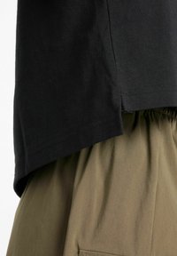 Dobladillo de camiseta negra parcialmente cubierto por pantalones oliva verde, plisados y ligeros; presentan textura suave de algodón y aberturas laterales para mayor movilidad.