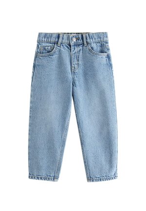 Smilende barn med krøllet hår står iført en almindelig hvid t-shirt, løse blå jeans og hvide sneakers, med hænderne i lommerne foran en hvid baggrund.