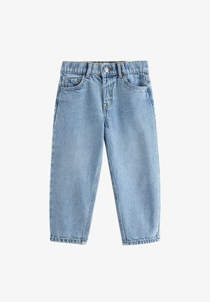 Smilende barn med krøllet hår står iført en almindelig hvid t-shirt, løse blå jeans og hvide sneakers, med hænderne i lommerne foran en hvid baggrund.