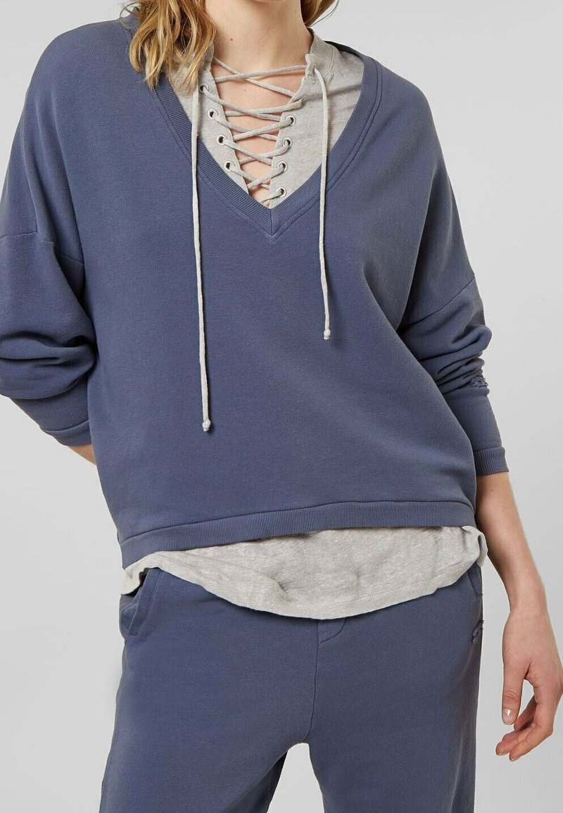 Přerostlá modrá mikina s výstřihem do V a šedou vrstvenou košilí, která má šněrovací detaily a stahovací šňůrky. Měkká látka s dlouhými rukávy.