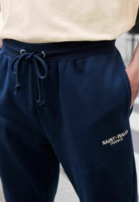 Pantalonii de trening bleumarin au un talie cu cordon de ajustare cu elemente metalice argintii, buzunare laterale și un logo brodat "SAINT-MALO FRANCE" în bej.