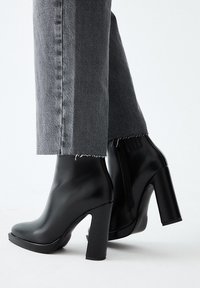 Botines negros de tacón alto con un acabado de cuero sintético suave, punta cuadrada y tacón grueso, combinados con unos vaqueros grises recortados con dobladillo deshilachado.