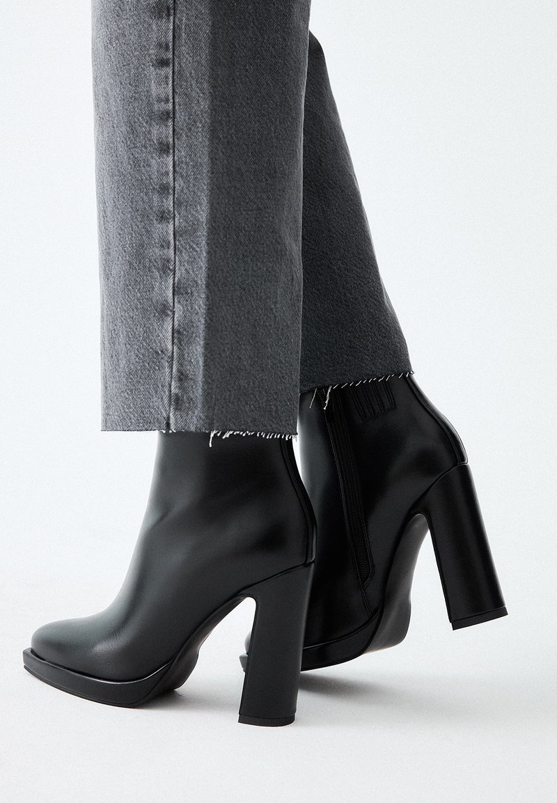 Botines negros de tacón alto con un acabado de cuero sintético suave, punta cuadrada y tacón grueso, combinados con unos vaqueros grises recortados con dobladillo deshilachado.