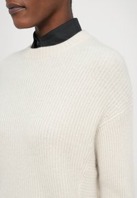 Crème ribgebreide trui met een ronde halslijn en verlaagde schouders, gelaagd over een zwarte blouse met kraag. Zachte textuur, minimalistisch ontwerp.
