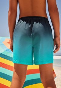 Shorts de bain à dégradé bleu avec une ceinture élastique, une texture lisse et une longueur short, présentés devant un parasol de plage coloré.
