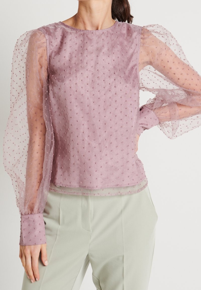 Femme portant un chemisier rose transparent à manches longues avec une texture à pois et des manches bouffantes, rentré dans un pantalon de couleur claire.