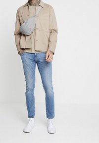 Veste légère beige portée par-dessus une chemise beige, associée à un jean bleu clair et des baskets blanches. Un sac bandoulière gris est porté en diagonale sur la poitrine.
