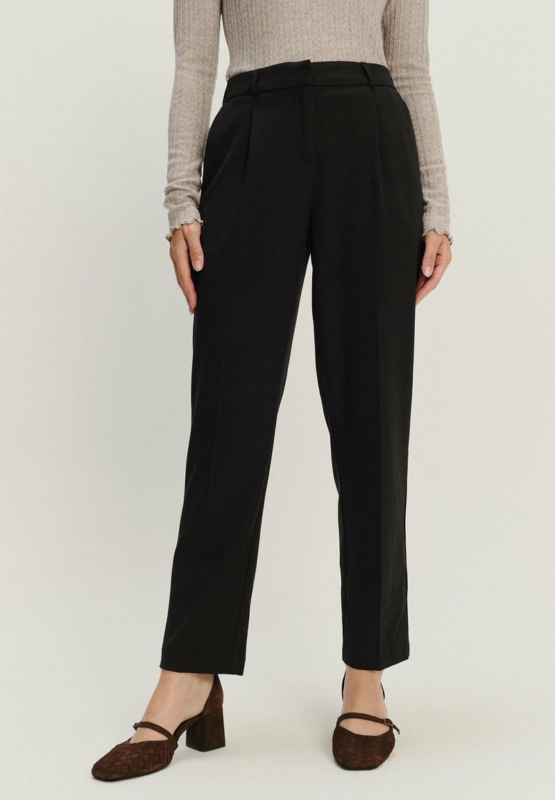 Schwarze, maßgeschneiderte Hose mit Falten, flachem Bund und hoher Taille. Kombiniert mit einem beigen, gerippten Langarmoberteil und braunen, hohen Schuhen.
