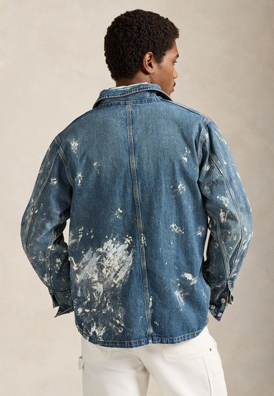 Άνδρας με την πλάτη γυρισμένη φοράει ένα μπλε τζιν μπουφάν με λευκές πιτσιλιές από μπογιά και λευκό παντελόνι, μπροστά σε απλό μπεζ φόντο.