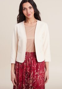 Witte blazer met driekwarts mouwen, open voorkant en twee zijzakken, gecombineerd met een beige top en een rode, gebloemde rok.