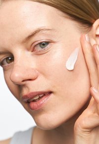 Ansiktsfuktighetskräm applicerad på kinden, vit krämig konsistens, slät yta, klar hudton, ljusreflektion, kort hår, naturlig makeup.