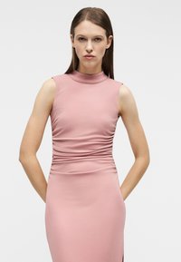 Γυναίκα με ίσια καστανά μαλλιά φοράει ροζ φόρεμα χωρίς μανίκια με ραφή, στέκεται με τα χέρια πίσω από την πλάτη, μπροστά σε απλό φόντο.