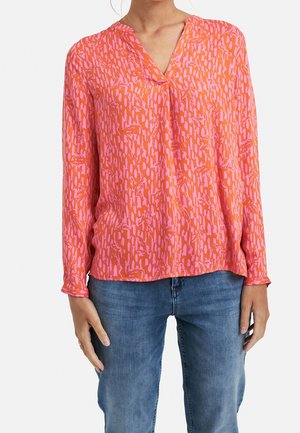 Femme portant un chemisier à manches longues, ample, avec des motifs abstraits roses et orange, associé à un jean bleu.