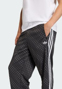 Czarne wzorzyste spodnie dresowe z motywem liści, białymi trzema paskami po bokach oraz kieszeniami. Tkanina wydaje się lekka i miękka.