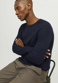 Pullover in maglia a coste di colore blu navy con scollo rotondo, dotato di maniche lunghe e una vestibilità comoda, abbinato a pantaloni color marrone chiaro.