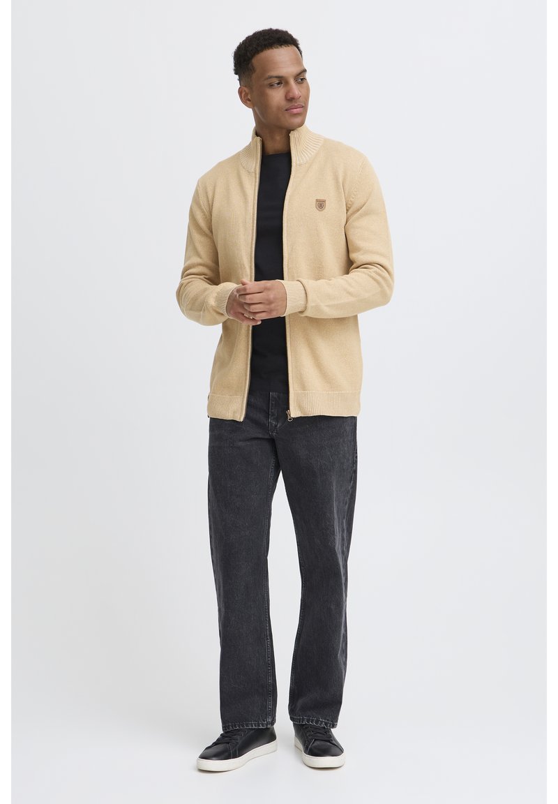 Cárdigan beige con cremallera y cuello alto, con puños y dobladillo acanalados, llevado sobre una camisa negra. El modelo lo combinó con jeans oscuros y zapatillas negras.