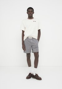 Ανοιχτόχρωμο T-shirt με κοντά μανίκια, κόκκινο γραφικό και ριμπ γιακά; σορτς με ασπρόμαυρο καρό; καφέ παπούτσια με λευκές κάλτσες.