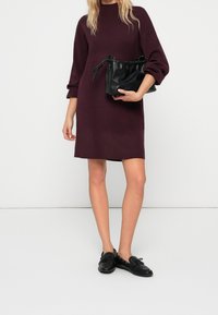 Robe en maille bordeaux avec manches jusqu'aux coudes, associée à un sac à cordon en cuir texturé noir et des chaussures plates noires.