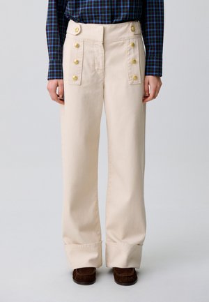 Personne portant un pantalon crème à jambes larges avec des boutons dorés, une chemise à carreaux bleue et des chaussures marron, debout sur un fond uni.