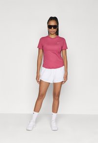 T-shirt de sport rose à manches courtes avec un col rond et le logo Nike, associé à un short blanc. Modèle portant des lunettes de soleil noires et des baskets blanches.