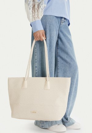Osoba nosící košili s krajkovými rukávy a širokými džíny drží velkou krémovou texturovanou tašku s pletenými držadly, stojící na bílém pozadí.