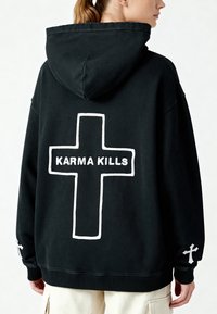 Felpa nera oversize con grafica di una croce bianca e il testo "KARMA KILLS" sulla parte posteriore, con polsini a costine e un tessuto testurizzato.