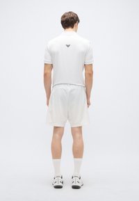 Biele polo tričko s čiernym logom na chrbte, v kombinácii s bielymi šortkami, bielymi ponožkami a čierno-bielymi športovými topánkami. Jednoduchý dizajn.