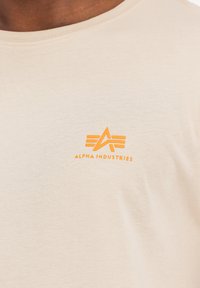 Beige Baumwoll-T-Shirt mit einem markanten orangefarbenen "ALPHA INDUSTRIES" Logo und einem stilisierten Flügel-Design auf der Vorderseite.