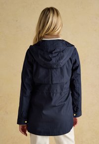 Frau mit langen blonden Haaren, die eine dunkelblaue Kapuzenjacke und weiße Hose trägt, steht vor einer beige strukturierten Wand.