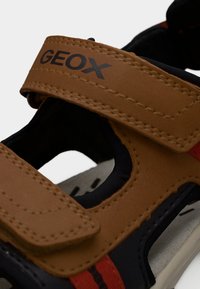 Καφέ δερμάτινα σανδάλια με μαύρες και κόκκινες λεπτομέρειες, ρυθμιζόμενους ιμάντες με βέλκρο, ανοίγματα για αερισμό και επενδυμένα πατάκια.