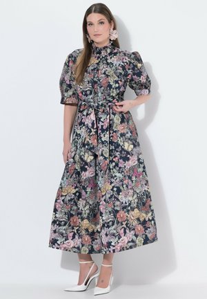 Frau trägt ein geblümtes, knöchellanges Kleid mit Puffärmeln, hohem Kragen, gebundener Taille und weißen Knöchelriemen-Absätzen, stehend vor weißem Hintergrund.