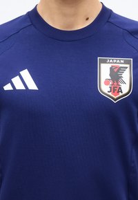 Μπλε αθλητική φανέλα με λευκό λογότυπο Adidas στην αριστερή πλευρά και σήμα της Ιαπωνικής Ποδοσφαιρικής Ομοσπονδίας στη δεξιά πλευρά του στήθους.