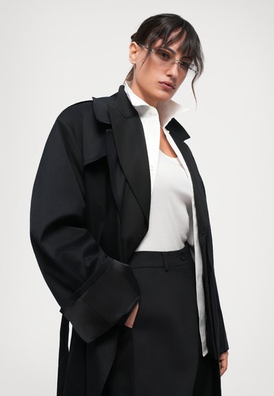 Svart oversize trenchcoat med vida ärmar över en vit skjorta och en fitted vit linne, kombinerat med svarta skräddarsydda byxor.