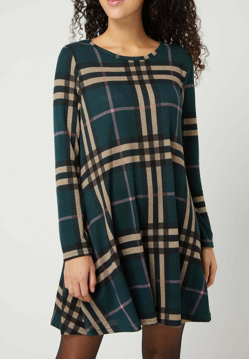 Femme portant une robe à manches longues à carreaux vert foncé, beige et noir avec un col rond, debout devant un fond uni.