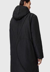Czarny ocieplany płaszcz z kapturem, z geometrycznymi wzorami przeszyć, długą sylwetką i bocznymi kieszeniami na zamek. Gładka tekstura.
