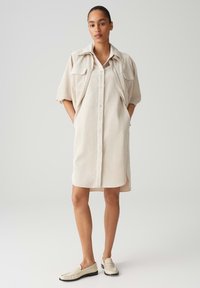 Helles beigefarbenes Hemdblusenkleid mit kurzen Ärmeln, Vorderknöpfen, zwei Brusttaschen und seitlichen Schlitzen. Kombiniert mit hellbeigen Slippern. Glatte Textur.