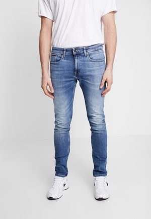 Man draagt blauwe slim-fit jeans, wit T-shirt en witte Nike sneakers, staand tegen een effen lichte achtergrond.
