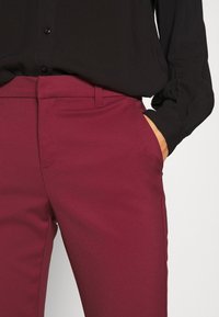 Pantalons taillés bordeaux avec une texture lisse, dotés de poches latérales et d'une coupe slim. Associés à une chemise noire à boutons.