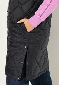 Černá prošívaná vesta se zipem vpředu a patentky. Obsahuje boční kapsy a růžovou košili s dlouhým rukávem pod ní. Viditelné strukturované tkaniny a švy.