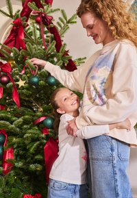 Ziemassvētku eglīte ar zaļām adatām, rotāta ar sarkaniem lentēm, zelta zvaigznēm un tirkīza rotājumiem. Sieviete gaišā sporta džemperī un džinsos apskauj meiteni baltā krekla.