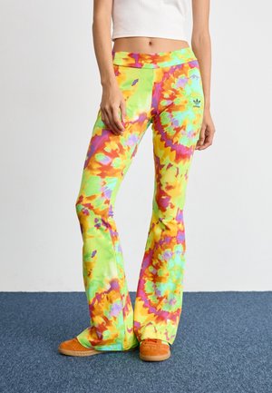 Osoba nosząca kolorowe rozkloszowane spodnie tie-dye i pomarańczowe buty, stojąca na niebieskim wykładzinowym podłodze przy prostej białej ścianie.