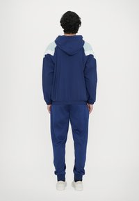 Navy tracksuit with a hood, featuring light blue accents on shoulders and cuffs. Smooth texture, elastic cuffs and waistband, with a relaxed fit.
