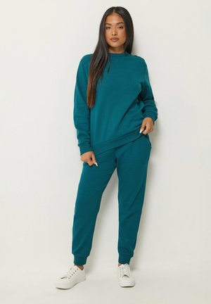 Donna con felpa a maniche lunghe verde acqua e pantaloni da jogging abbinati con scarpe da ginnastica bianche, in piedi contro un muro chiaro e semplice.