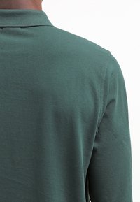 Tmavě zelená polo košile s dlouhými rukávy, vyrobená z texturované tkaniny a s klasickým límcem. Pohled zezadu ukazuje hladký a jednoduchý design.