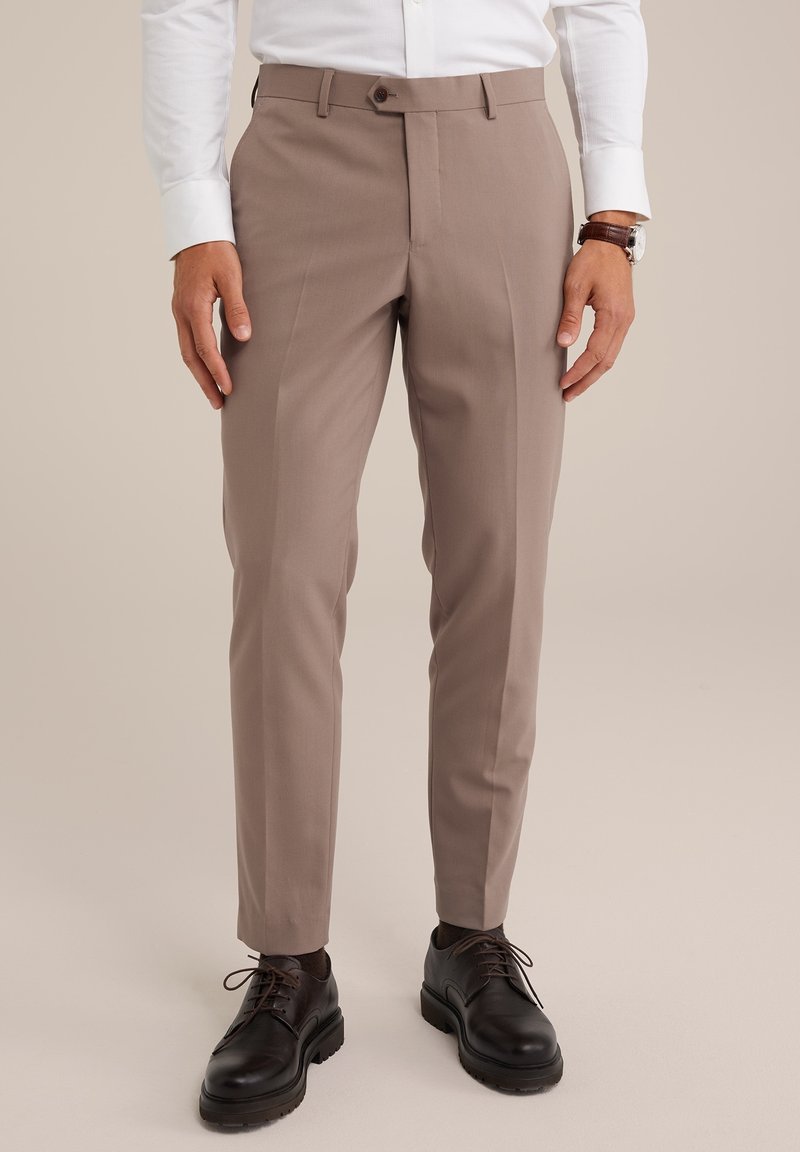 Homme portant un pantalon beige slim fit, une chemise blanche, une montre en cuir marron et des chaussures noires à lacets sur un fond neutre.