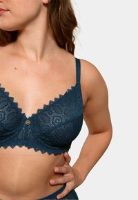 Soutien-gorge en dentelle teal avec des bords festonnés, présentant des motifs géométriques, des bretelles ajustables et un bouton décoratif central.